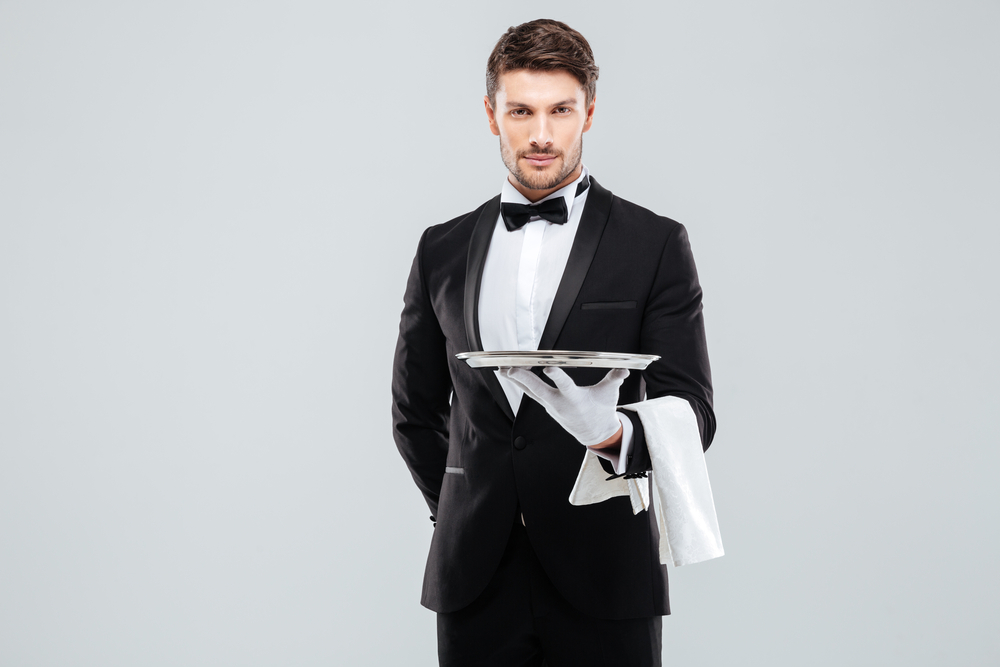 Handsome yong waiter in tuxedo and gloves holding empty tray and napkin
