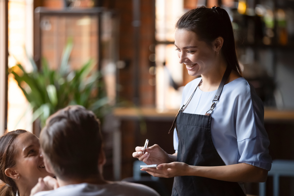 Smiling female waitress take order talk to clients cafe restaurant visitors couple, friendly professional woman server wear apron write dinner food menu choice, serving staff good customer service